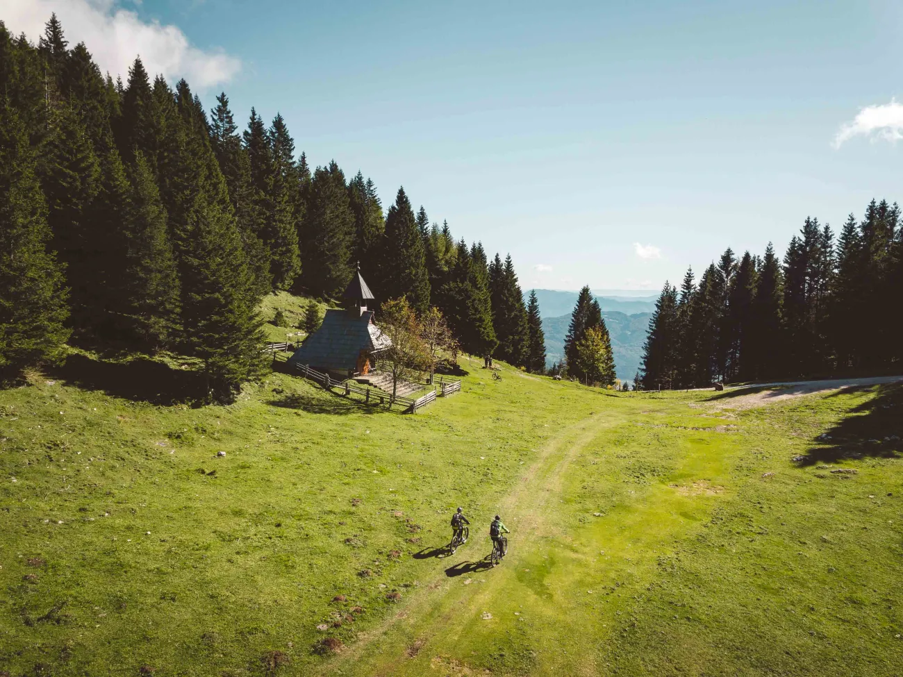 Gorska jasa, kapelica in gozdna pokrajina s kolesarji v daljavi